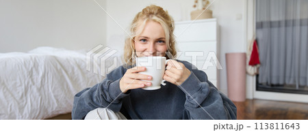 Image of young teenage girl sitting in her bedroom on floor, drinking cup of tea and enjoying day at home, smiling and looking at camera 113811643