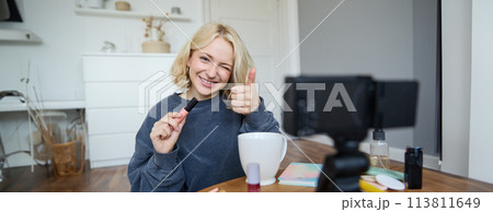 Portrait of smiling, beautiful young woman, lifestyle blogger recording a video in her room, sitting in front of camera on stabiliser, showing thumbs up and wink face, recommending mascara 113811649