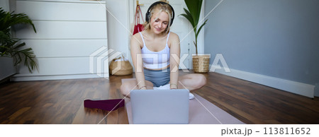 Portrait of smiling sporty woman at home, doing workout on rubber yoga mat, using laptop and wireless headphones Portrait of smiling sporty woman at home, doing workout on rubber yoga mat, using laptop and wireless headphones 113811652