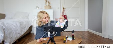 Portrait of smiling blond woman, teenage girl records a vlog on video camera, vlogger shows eyeshadows, tutorial how to put makeup on, sits in her room on floor near coffee table Portrait of smiling blond woman, teenage girl records a vlog on video camera, vlogger shows eyeshadows, tutorial how to put makeup on, sits in her room on floor near coffee table 113811658
