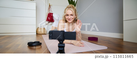 Portrait of smiling blond woman saying hello to her followers, fitness instructor recording video of herself showing workout exercises at home, lying on rubber yoga mat, looking at digital camera 113811665