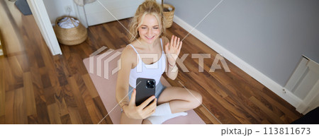 Portrait of fit, sporty young fitness instructor, woman gives online workout sessions to clients, waves hand at smartphone, sits on yoga mat and shows exercises Portrait of fit, sporty young fitness instructor, woman gives online workout sessions to clients, waves hand at smartphone, sits on yoga mat and shows exercises 113811673