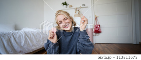Portrait of young woman, content creator, showing beauty makeup products at camera, sitting on floor in bedroom and smiling Portrait of young woman, content creator, showing beauty makeup products at camera, sitting on floor in bedroom and smiling 113811675