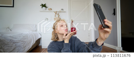 Image of young woman sits on floor in her bedroom, takes selfies on smartphone, sends air kisses at mobile phone camera, video chats online using app Image of young woman sits on floor in her bedroom, takes selfies on smartphone, sends air kisses at mobile phone camera, video chats online using app 113811676