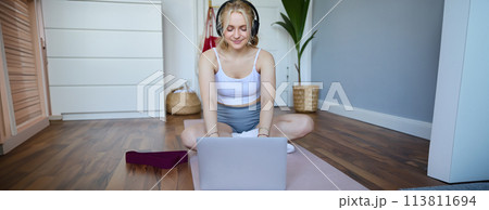 Portrait of young sporty woman following online video instructions during fitness workout, using laptop, sitting on yoga mat 113811694