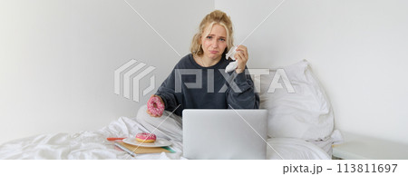 Portrait of woman watching sad movie on laptop, crying and wiping tears off, eating doughnut and drinking tea 113811697