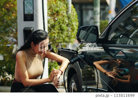 Smiling woman checking time on wristwatch when waiting for her electric car to fully charge 113811699