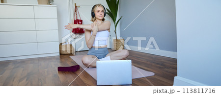 Wellbeing and fitness concept. Woman in headphones, sits on yoga mat, watches workout video on laptop, stretches arms, does exercises Wellbeing and fitness concept. Woman in headphones, sits on yoga mat, watches workout video on laptop, stretches arms, does exercises 113811716