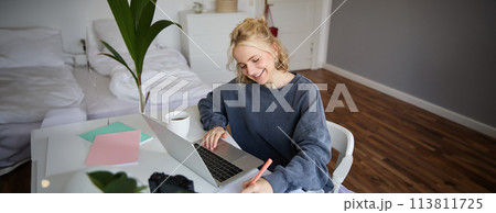 Portrait of smiling, beautiful blond woman, writing down notes, doing homework, studying from home, doing distance learning, online course, working remotely in her room 113811725