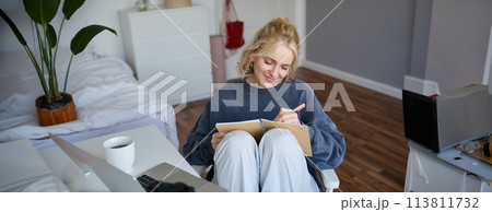 Portrait of young woman writing in diary, making notes in notebook, sitting on chair in a room in front of laptop, student doing homework 113811732