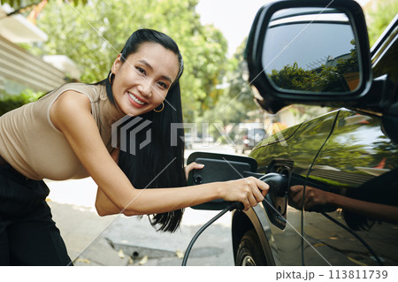 Happy woman inserting plug in electric car to charge battery Happy woman inserting plug in electric car to charge battery 113811739