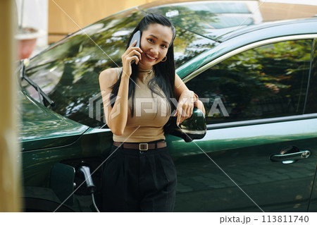 Smiling successful businesswoman standing next to electric car when talking on phone Smiling successful businesswoman standing next to electric car when talking on phone 113811740