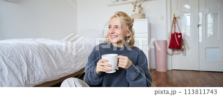 Lifestyle portrait of young woman sitting on bedroom floor with cup of tea, drinking from big white mug and looking aside, smiling happily 113811743