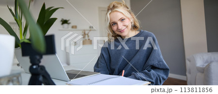 Portrait of beautiful smiling woman, student studying at home, remote education concept, sits in a room with laptop and writes down notes, uses her journal Portrait of beautiful smiling woman, student studying at home, remote education concept, sits in a room with laptop and writes down notes, uses her journal 113811856