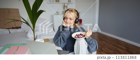 Image of happy woman sitting in a room, watching interesting tv show or movie on laptop, using screaming service, wearing headphones, eating dessert and drinking tea Image of happy woman sitting in a room, watching interesting tv show or movie on laptop, using screaming service, wearing headphones, eating dessert and drinking tea 113811868