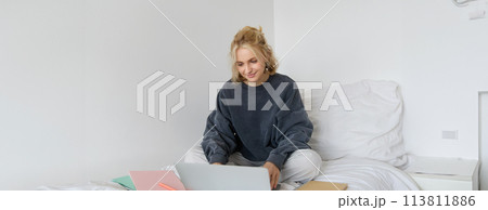 Image of happy young woman, student e-learning from home, connect to online course on her laptop, sits on bed with notebooks, smiling and looking happy Image of happy young woman, student e-learning from home, connect to online course on her laptop, sits on bed with notebooks, smiling and looking happy 113811886