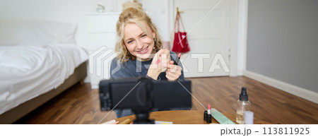 Image of young stylish woman, blogger recording a beauty lifestyle video of her picking best lipstick, showing lip balm swatches on her skin, sitting in front of digital camera in empty room Image of young stylish woman, blogger recording a beauty lifestyle video of her picking best lipstick, showing lip balm swatches on her skin, sitting in front of digital camera in empty room 113811925