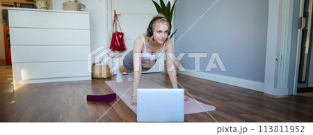 Portrait of young athletic woman in wireless headphones, standing in plank, following online workout video, using laptop to join training session on the internet 113811952