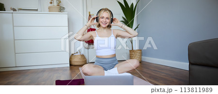 Portrait of fit and healthy young woman in wireless headphones, does workout, using online video tutorials, listens to fitness instructor on laptop, exercising at home Portrait of fit and healthy young woman in wireless headphones, does workout, using online video tutorials, listens to fitness instructor on laptop, exercising at home 113811989