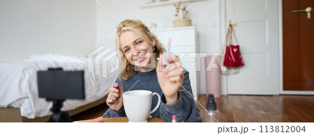 Portrait of smiling woman shows lip gloss, advertising makeup on her social media account, recording video on digital camera, content maker working on new project in her room Portrait of smiling woman shows lip gloss, advertising makeup on her social media account, recording video on digital camera, content maker working on new project in her room 113812004