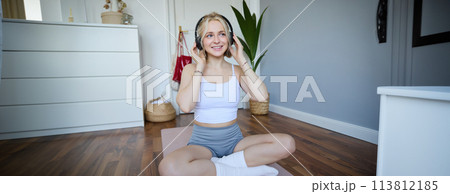 Young and sporty woman working out at home, doing fitness training alone on yoga mat, listens to music in wireless headphones 113812185