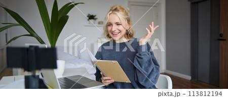 Portrait of young smiling blond woman, working from home, online chatting, using digital video camera, recording vlog, holding notebook, reading notes, explaining something 113812194