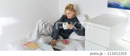Portrait of young woman, student studying in her bed, relaxing while preparing homework, eating doughnut, using laptop in bedroom and drinking tea Portrait of young woman, student studying in her bed, relaxing while preparing homework, eating doughnut, using laptop in bedroom and drinking tea 113812203
