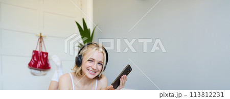 Vertical shot of young fitness woman in headphones, lying on rubber mat, relaxing after workout training session, holding smartphone, smiling at camera Vertical shot of young fitness woman in headphones, lying on rubber mat, relaxing after workout training session, holding smartphone, smiling at camera 113812231