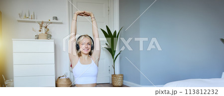 Portrait of woman doing fitness exercises at home, watches sports video on laptop, wearing wireless headphones, follows workout instructions, sits on rubber mat in her room 113812232