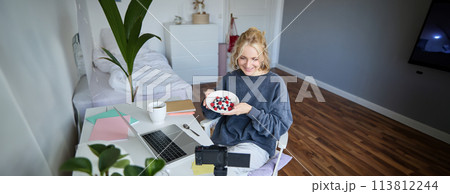 Portrait of cute young girl vlogger, showing her breakfast on camera, recording vlog in her bedroom, holding dessert, eating 113812244