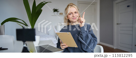Portrait of young creative woman, content maker, sitting in room, working from home, using laptop, holding notebook, raising finger, has an idea, eureka gesture 113812295
