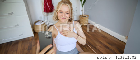 Young sporty fitness trainer, yoga instructor does live stream, takes selfies on smartphone while sitting on rubber mat at home, recording her workout training session Young sporty fitness trainer, yoga instructor does live stream, takes selfies on smartphone while sitting on rubber mat at home, recording her workout training session 113812382