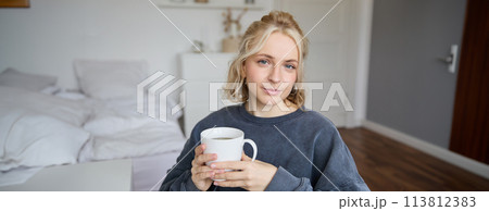Close up portrait of smiling young blond woman, sits with cup of tea in bedroom, rests at home, enjoys her coffee Close up portrait of smiling young blond woman, sits with cup of tea in bedroom, rests at home, enjoys her coffee 113812383