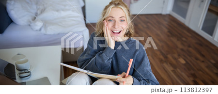 Portrait of happy, young blond woman, sitting in bedroom with notebook and pen, laughing and smiling, writing in journal, doing homework and smiling 113812397