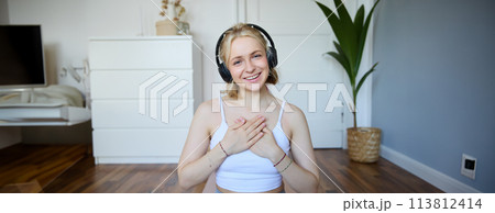 Portrait of young happy woman in headphones, looking with relaxed, friendly face expression, holding hands on her chest and smiling 113812414