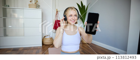 Portrait of fit and healthy woman workout at home, wearing headphones, showing smartphone screen, empty dark display 113812493