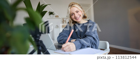 Portrait of young woman, lifestyle blogger, recording video of herself, making notes, writing in journal, sitting in front of laptop in a room and studying Portrait of young woman, lifestyle blogger, recording video of herself, making notes, writing in journal, sitting in front of laptop in a room and studying 113812544