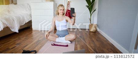 Portrait of stylish young woman, sitting on rubber mat on floor at home, taking selfie while doing workout, yoga session, posting photo on social media Portrait of stylish young woman, sitting on rubber mat on floor at home, taking selfie while doing workout, yoga session, posting photo on social media 113812572