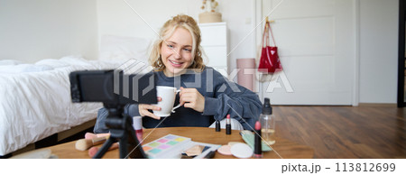 Blogging, modern technology, e-commerce and people concept. Happy smiling woman beauty video blogger holding cup of tea applying makeup while recording beauty tutorial at home for her video blog 113812699