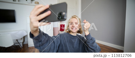 Portrait of young stylish girl sits on bedroom floor, takes selfies on her smartphone, posing for photo on social media app, smiling and looking happy at camera Portrait of young stylish girl sits on bedroom floor, takes selfies on her smartphone, posing for photo on social media app, smiling and looking happy at camera 113812706