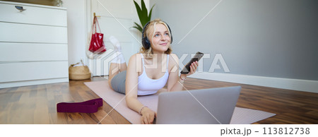 Portrait of happy, smiling sporty woman, doing workout at home, using laptop to watch workout videos in headphones, checking sports app on mobile phone 113812738
