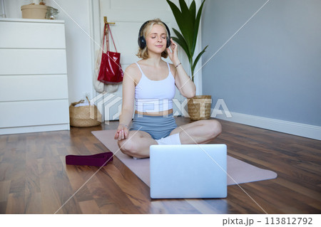 Portrait of fit and healthy woman at home, practice yoga, sitting on rubber mat, listening to instructions online, using meditation music to relax, following guidance on laptop Portrait of fit and healthy woman at home, practice yoga, sitting on rubber mat, listening to instructions online, using meditation music to relax, following guidance on laptop 113812792