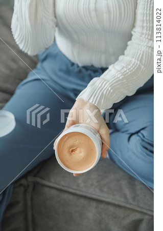 A woman sits on the couch holding jar with cosmetic cream 113814022