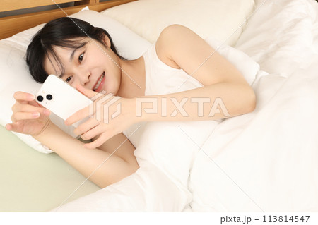 ベッドで寝ながらスマホを見る笑顔若い女性 113814547