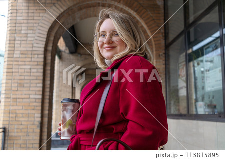 Young plus size woman in burgundy coat with cup of coffee walking in city. Portrait against background of an architectural arch 113815895