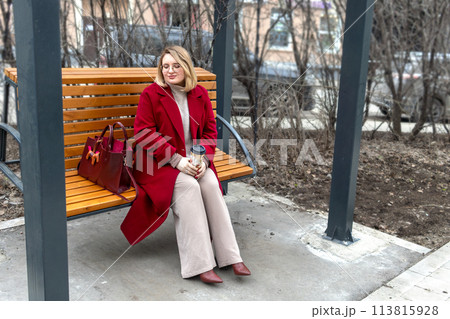 Urban loneliness: full woman in red coat and with coffee in hand on city swing in sad reverie 113815928