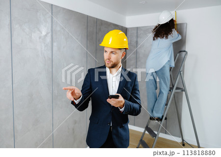 A young businessman holds a phone in his hands and checks the work of a designer in creating an interior in a room. The concept of repair on a modern design. 113818880