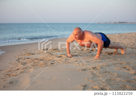 An elderly man is pushing up on the sea coast. Sports at sea middle-aged men. Healthy lifestyle on vacation. An elderly man is pushing up on the sea coast. Sports at sea middle-aged men. Healthy lifestyle on vacation. 113822250