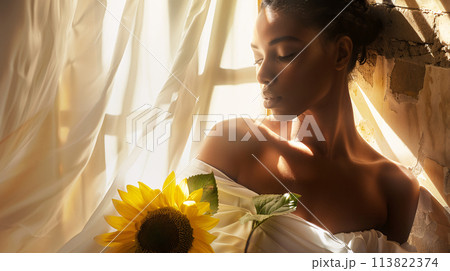 woman with sun-kissed skin draped in a flowing white dress, holding a single sunflower 113822374