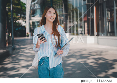 A woman is walking down the street holding a cell phone and a laptop 113822881